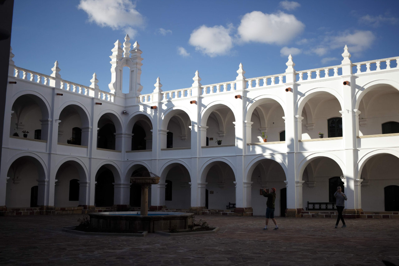 The interior courtyard of a colonial-style building featuring two stories of white arches and balconies under a blue sky; in the center stands a stone fountain basin on a paved ground