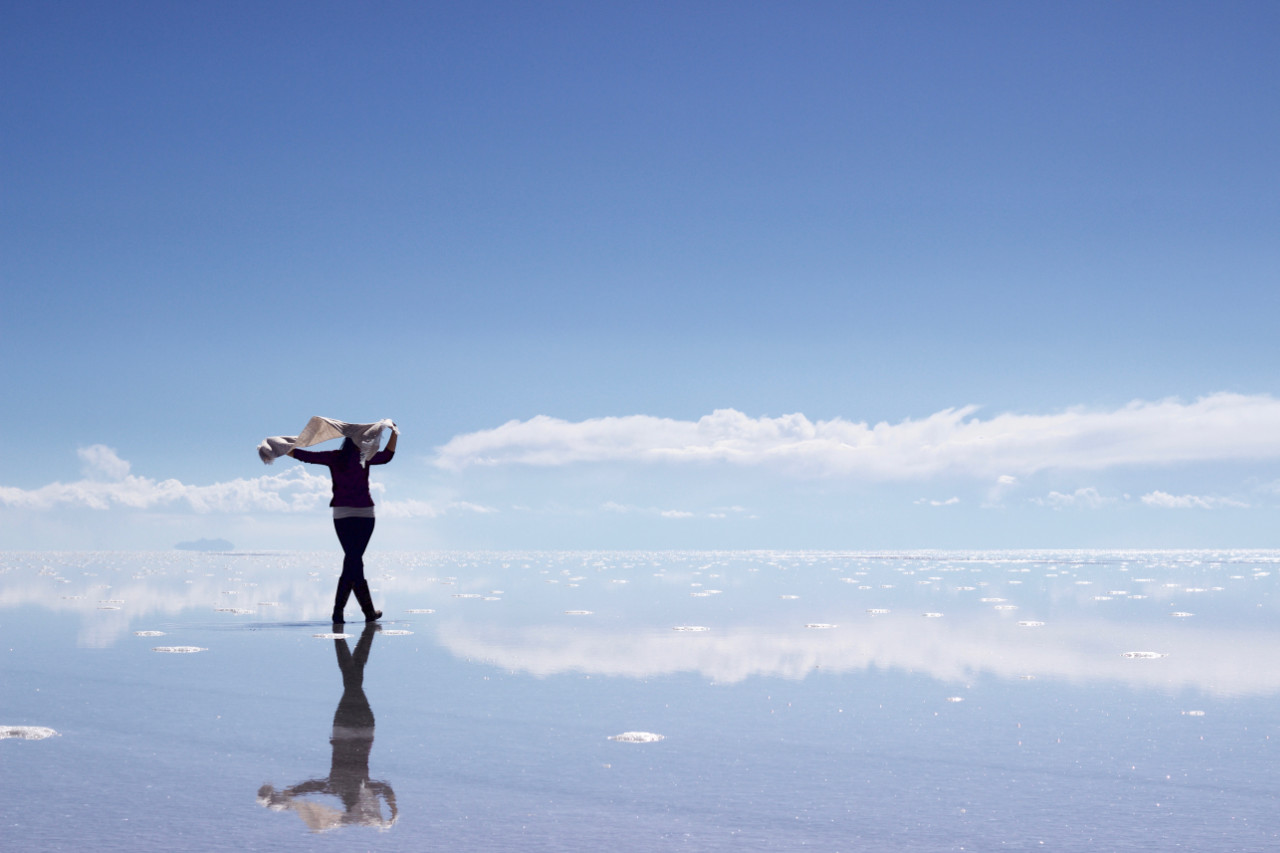A woman standing on the edge of a vast salt flat that acts like a mirror reflecting the bright blue sky above. They are holding a white cloth up to their head while looking out at the horizon where clouds meet the water, with small mounds of salt crystals visible around them.