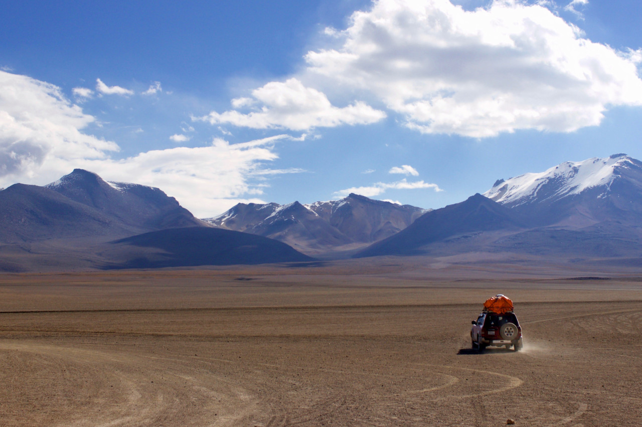 An off-road vehicle driving away across a vast, flat desert plain towards distant mountains; the car carries an orange bundle on its roof rack and kicks up dust behind it beneath a bright blue sky filled with scattered white clouds.