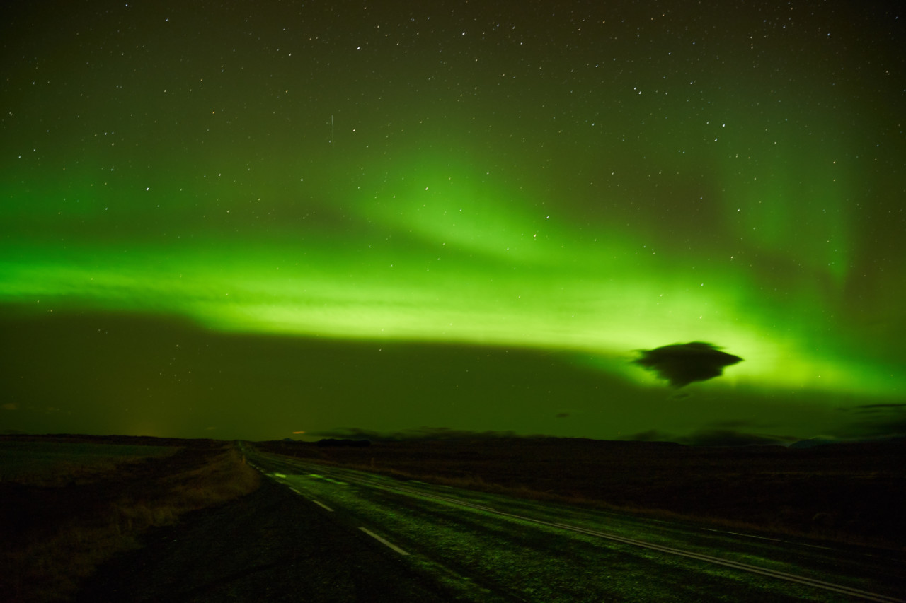 A night landscape capturing the vibrant green glow of the aurora borealis dancing across a starry sky above a dark, empty road that stretches from the bottom left into the distance through flat terrain.