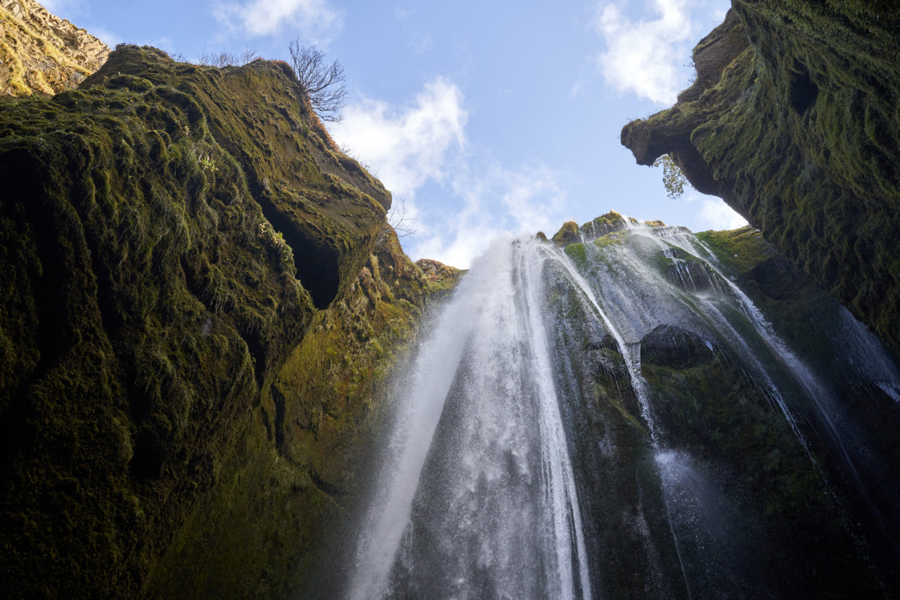 A low-angle perspective looking straight up at a powerful waterfall cascading between steep cliffs covered in thick, textured green moss, framing patches of blue sky and white clouds visible through the gap at the top of the gorge.