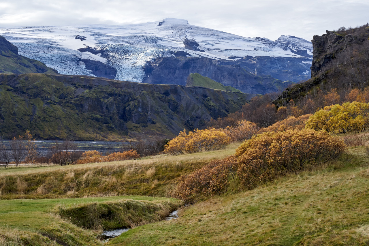 A scenic valley view dominated by a massive glacier covering the mountain peak in the background; below it are dark rocky slopes leading down to rolling hillsides dotted with yellow autumn bushes and a small stream flowing near a body of water on the left.